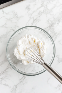 This image shows a medium-sized bowl where heavy cream, powdered sugar, and vanilla extract are being whisked together, forming soft peaks that will be used to top the refreshing raspberry Italian soda.