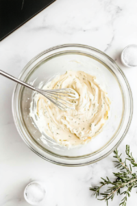 This image shows a large mixing bowl with vegan mayo, apple cider vinegar, sugar, celery seed, and salt being whisked together to create a smooth and tangy dressing for the vegan coleslaw.