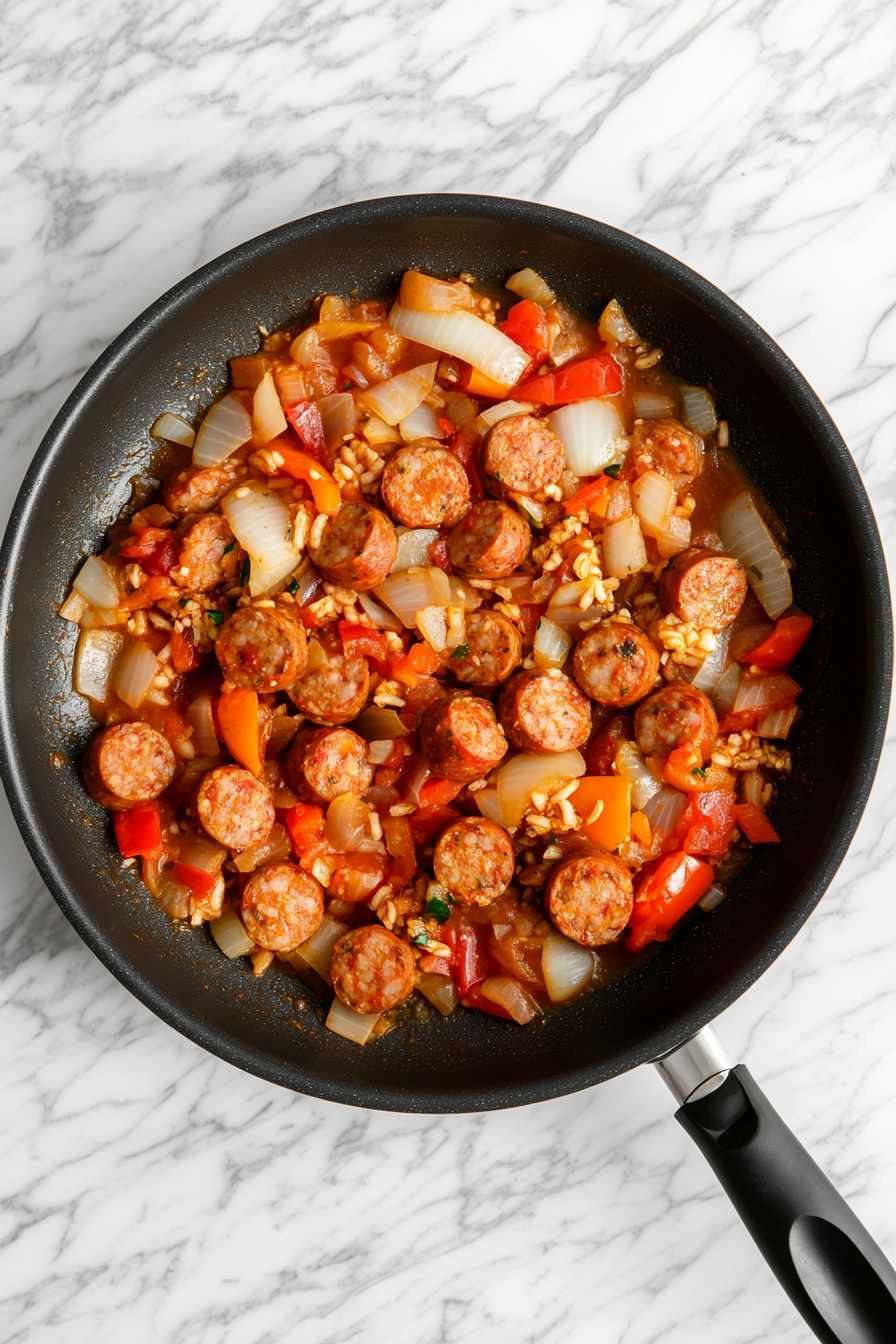 This image shows rice, chicken broth, tomato paste, and a blend of spices being added to the skillet with sausage and vegetables, preparing the ingredients to simmer together in the smoked sausage and rice dish.