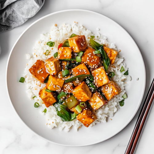 This image shows a bowl of Chinese tofu stir fry with golden fried tofu cubes, sautéed vegetables, and a savory sauce, served over steamed rice and garnished with fresh green onions and sesame seeds for a delicious meal.