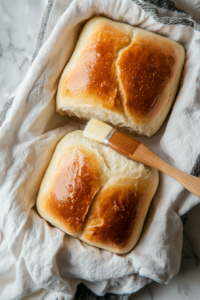 This image shows freshly baked bread loaves being brushed with water and wrapped in a tea towel to keep the crust soft and tender