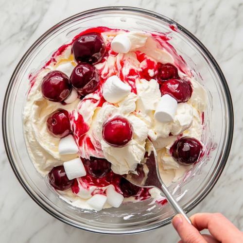 This image shows a serving of cherry cheesecake fluff, featuring a smooth and fluffy cheesecake filling mixed with cherry pie filling and mini marshmallows, creating a light and sweet dessert perfect for any occasion.