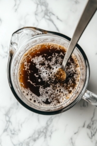 This image shows cold water, granulated sugar, and root beer extract being mixed together in a large pitcher to create the base for homemade root beer.