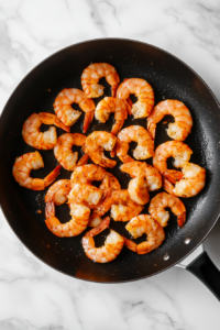 This image shows the seasoned shrimp being cooked in a hot skillet with avocado oil, turning opaque and curling up as they cook.