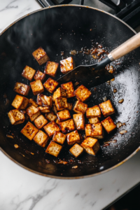 This image shows cubed firm tofu sizzling in a hot skillet, frying until it turns golden brown on all sides, which will give the tofu a crispy texture for the stir fry.