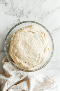 This image shows bread dough placed in a large bowl and covered with a damp tea towel, allowing it to rise in a warm spot until it doubles in size.