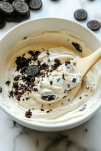 This image shows chopped Oreos being gently folded into the creamy cheesecake mixture, which is then spread evenly over the cookie dough and Oreo layer in the baking pan.