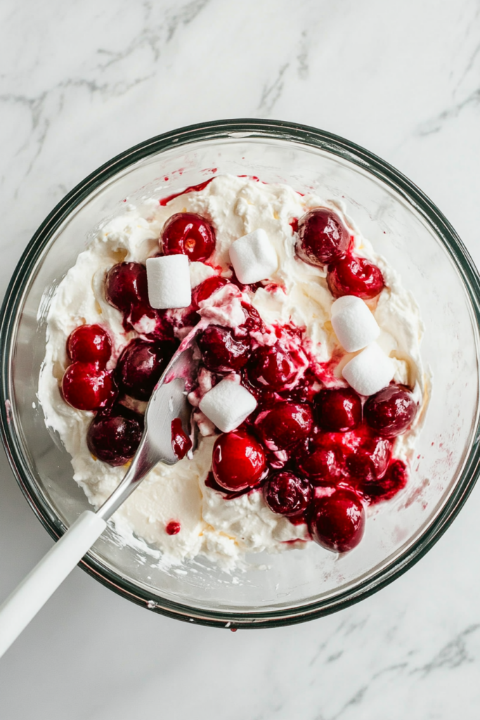 This image shows cherry pie filling and mini marshmallows being gently folded into the cheesecake mixture, giving the dessert its signature sweet and tangy cherry flavor with a fluffy texture.