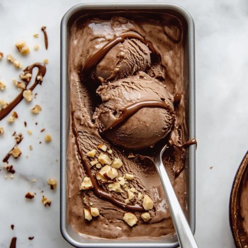 This image shows a baking pan of rich, velvety homemade Nutella ice cream, beautifully garnished with chopped hazelnuts and a drizzle of chocolate sauce, ready to be enjoyed.