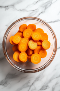 This image shows sliced carrots placed in a microwave-safe bowl with a small amount of water. The bowl is covered and being microwaved to soften the carrots for the vegan chicken noodle soup.