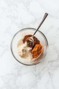 This image shows the process of mixing kosher salt, black pepper, onion powder, garlic powder, and smoked paprika in a small bowl to create a flavorful spice rub for baked chicken.