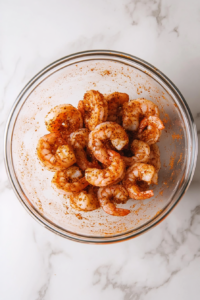 This image shows shrimp being evenly coated with a seasoning mixture of paprika, garlic powder, cayenne pepper, salt, and black pepper in a bowl.