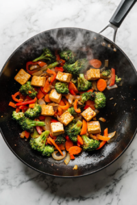 This image shows a colorful mix of julienned onions, bell peppers, carrots, and chopped broccoli being sautéed in a wok, adding flavor and texture to the tofu stir fry.