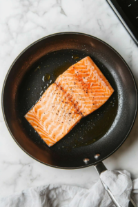 This image shows salmon fillets seasoned with salt and pepper, being seared in a skillet until they are golden brown and cooked through, forming the savory protein base for the spinach salmon pasta dish.