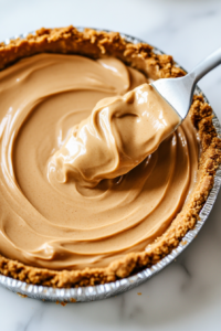 This image shows the smooth peanut butter filling being spread evenly into the graham cracker crust, assembling the pie.