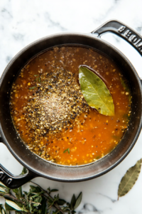 This image shows Italian seasoning being sprinkled into the pot, along with a bay leaf being added. The seasonings are enhancing the flavor of the vegan chicken noodle soup.