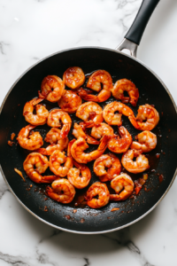 This image shows cooked shrimp being tossed in spicy Sriracha sauce in a skillet, ensuring they are evenly coated with the sauce.