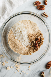 This image shows flour, baking soda, brown sugar, oats, coconut, pecans, and dried cherries being whisked together in a separate bowl, creating the dry ingredient mix for the breakfast muffins.