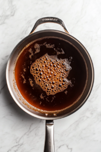 A top-down view of the skillet with broth being poured in. The broth is simmering and reducing by half, thickening the mixture. The scene captures the reduction process with the liquid bubbling in the skillet.