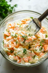 Top-down view of mayonnaise being spooned into the mixing bowl with the salad ingredients, with freshly chopped dill sprinkled on top. A spatula is prepared to fold the ingredients together.