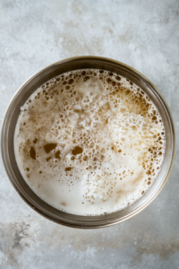 Top-down view of the proofed yeast mixture in the mixing bowl, with ¼ cup of vegetable oil and 1 ½ teaspoons of salt being added. The oil is slightly pooling on the surface, ready to be incorporated.