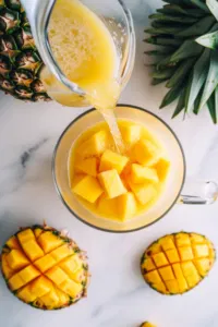 Top-down view of chilled Dole Pineapple juice and mango nectar being poured into the pitcher with the lemonade base, on a clean countertop.