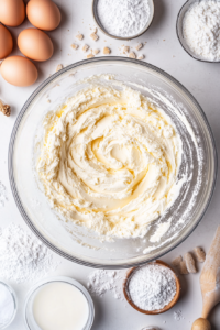 Top-down view of the same mixing bowl as powdered sugar, milk, and vanilla extract are gradually mixed into the cream cheese and butter. The frosting is forming a smooth consistency, with a few visible granules of powdered sugar.