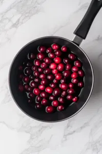 Top-down view of cranberries gently added to the cooled sugar syrup in the saucepan. The cranberries are fully submerged, soaking in the sweet mixture, preparing to absorb the syrup for 10-15 minutes.