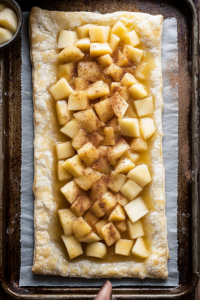 Top-down view of puff pastry on a baking sheet with 8 ounces of apple pie filling spooned down the center. Ground cinnamon is sprinkled over the filling, ready for folding.