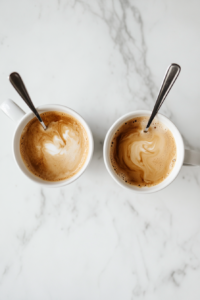 Top-down view of the two mugs of coffee on the white marble cooktop. Irish cream liqueur, hazelnut liqueur, and Irish whiskey are being poured into each mug and stirred with a spoon, highlighting the smooth blending of the liqueurs with the coffee.