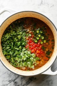 A top-down view of the pot as chicken stock is being poured in, along with chopped jalapeños (or green chilies), fresh cilantro, ground cumin, and dried oregano. The mixture is simmering, with bubbles forming on the surface. The scene captures the colorful ingredients melding together in the broth.