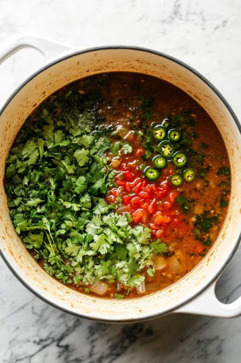 A top-down view of the pot as chicken stock is being poured in, along with chopped jalapeños (or green chilies), fresh cilantro, ground cumin, and dried oregano. The mixture is simmering, with bubbles forming on the surface. The scene captures the colorful ingredients melding together in the broth.