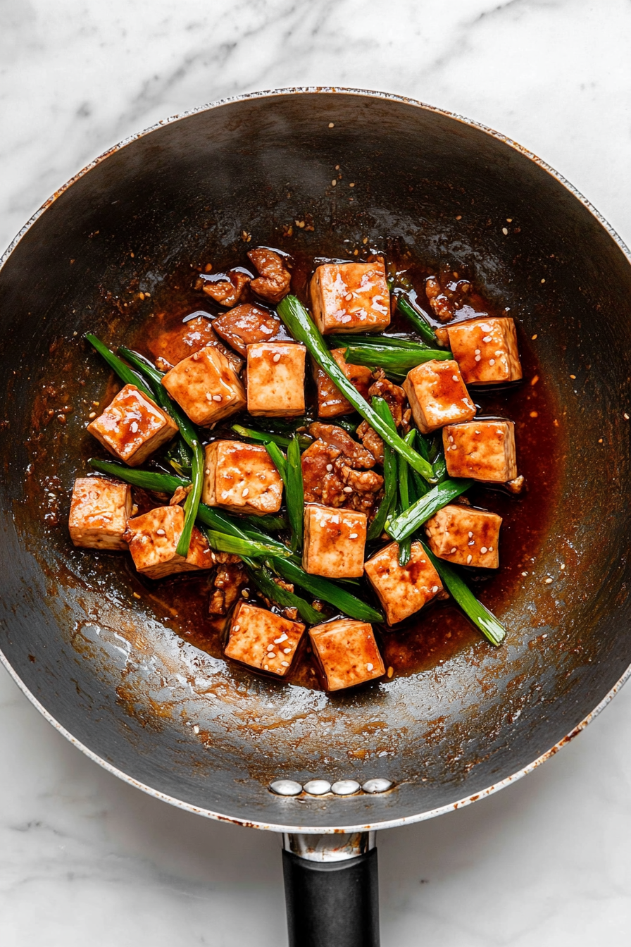 A top-down view of a wok on a white marble cooktop. Pan-fried tofu slices and the green parts of scallions are being added to the sauce and cooked meat, stir-frying quickly to combine.