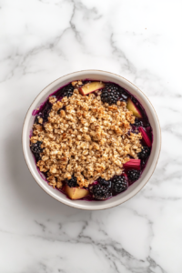 Top-down view of a greased 9x13-inch baking dish on the white marble cooktop. The fruit mixture is evenly spread in the dish. Clumps of crumble dough are dropped over the fruit, with chopped walnuts and melted butter drizzled on top. The scene shows the layered assembly of the crumble.