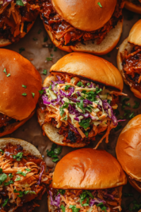 Top-down view of BBQ jackfruit-tofu mixture being spooned onto toasted burger buns, topped with pineapple coleslaw or simple slaw. The sandwich is colorful and hearty, ready to serve with rich barbecue and refreshing slaw.