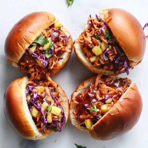 Top-down view of BBQ jackfruit-tofu mixture being spooned onto toasted burger buns, topped with pineapple coleslaw or simple slaw. The sandwich is colorful and hearty, ready to serve with rich barbecue and refreshing slaw.