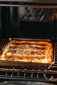 Top-down view of the baking pan inside the oven, with the cream cheese bars baking at 350°F (or 325°F for a dark pan). The top layer is golden and slightly puffed as the bars bake for 35 minutes.