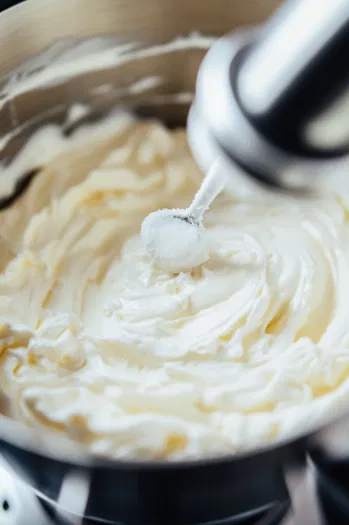 Top-down view of egg whites being whipped in a stand mixer, with soft peaks forming. A tablespoon of sugar is ready to be added gradually.