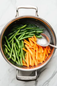 Top-down view of green beans and matchstick-sized carrots being blanched in a large pot of salted boiling water on a white marble cooktop, with a slotted spoon ready to scoop them into an ice water bath nearby.