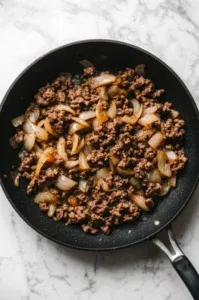 This image shows ground beef being added to the caramelized onions in the skillet, browning until fully cooked and seasoned with mustard, chicken bouillon, Sazon, garlic powder, onion powder, salt, and pepper.