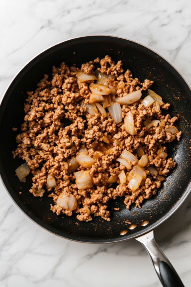 A top-down view of a large skillet on the white marble cooktop. Ground turkey is being broken up and browned in 1 tablespoon of avocado oil over medium-high heat. Minced garlic and diced onions are added as the turkey starts to cook, turning golden while the garlic and onions soften