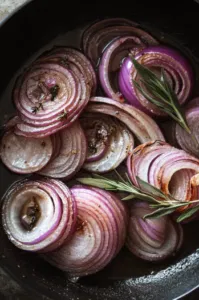 Top-down view of sliced onions in a skillet slowly caramelizing in butter and olive oil. Soy sauce, rosemary, thyme, and sage are sprinkled over the onions as they cook, with the onions being stirred occasionally for even caramelization.