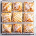 Top-down view of cooled cheese danishes on a wire rack over a white marble cooktop. A smooth glaze made from powdered sugar, vanilla extract, and milk is being drizzled over each danish, focusing on the shiny glaze cascading over the golden pastries.