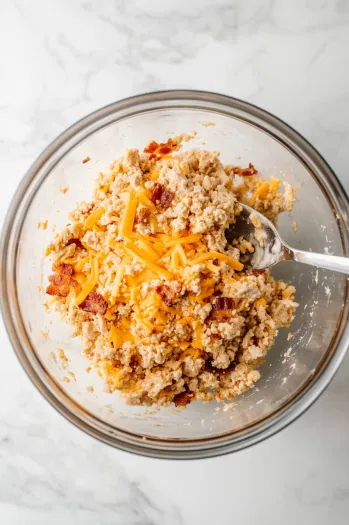 This image shows ground chicken being mixed with egg, breadcrumbs, ranch dressing mix, shredded cheddar cheese, and chopped bacon in a large bowl.