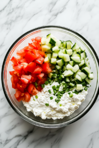 A top-down view of a medium mixing bowl on the white marble countertop. In the bowl, 16 ounces of drained cottage cheese, the chopped tomatoes, sliced green onions, and diced cucumbers are being added. The scene captures all ingredients coming together in the bowl, ready for mixing.