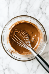 A top-down view of a mixing bowl on a clean kitchen countertop with a white marble cooktop background. Inside the bowl, milk, rum cream liqueur, and instant chocolate pudding mix are being combined. A whisk is mixing the ingredients, with the mixture starting to thicken. The focus is on the creamy consistency of the pudding mixture.