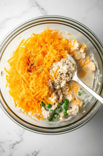 Top-down view of a large mixing bowl on a white marble countertop. Inside the bowl, drained tuna, cream of mushroom soup, milk, shredded cheddar cheese, frozen peas, black pepper, garlic powder, and onion powder are being mixed together with a large spoon.