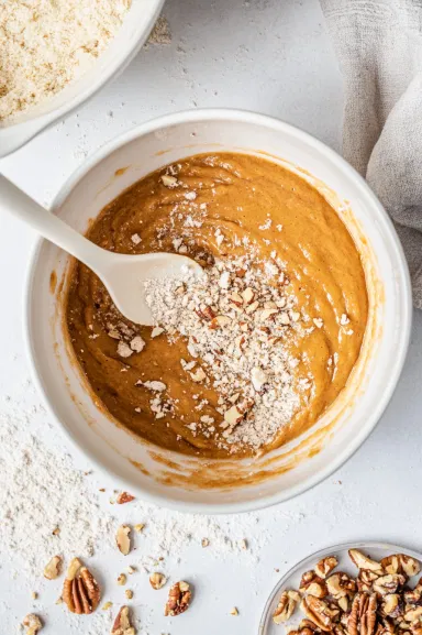 Top-down view of the wet banana mixture being poured into the bowl of dry ingredients. The ingredients are gently stirred together, forming a slightly lumpy batter, with chopped nuts being folded in, ready to be transferred to the pan.