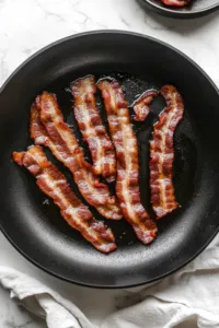 This image shows bacon being cooked in a large skillet over low heat to achieve crispy results. The bacon is then set aside with the drippings reserved.