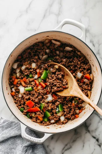 Top-down view of ground beef browning in a large pot on a white marble countertop, with chopped onions, bell peppers, jalapeño, and garlic being stirred in with a wooden spoon.